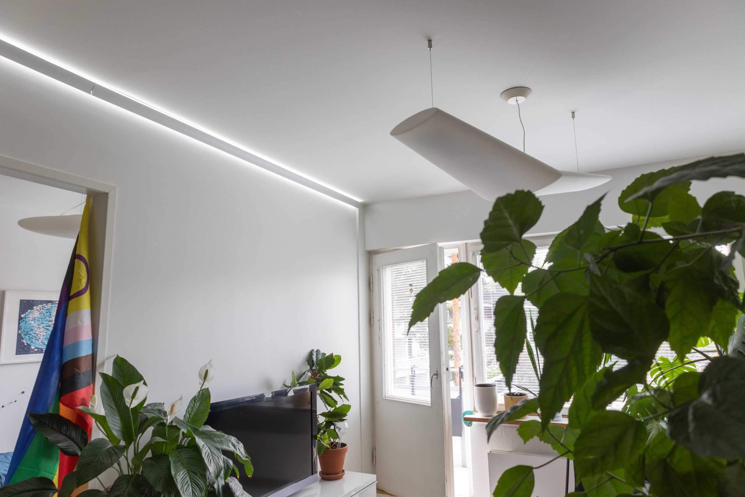 Moderni olohuone minimalistisella designilla sisältää suuria viherkasveja, tv ja värikkään lipun. Tyylikäs led-valaisin tarjoaa katossa eleganttien LED-elementtien rinnalla, kun luonnonvalo tulvii sisään ulos johtavista lasiovista.