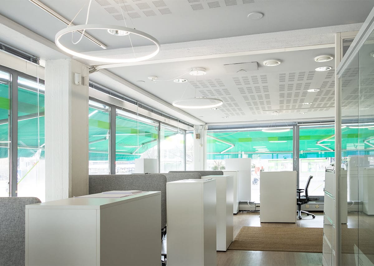 Moderni toimisto, jossa on valkoiset työpöydät, harmaat tuolit, pyöreät LED-valot ja suuret ikkunat vihreillä sävyillä. Vaaleat puulattiat ja matot lisäävät tilaa.