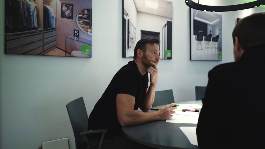 Mies, jolla on parta pöydässä, leuka kädellä, modernissa huoneessa; LED-valaistut julisteet seinillä esittelevät kaappimalleja. LED-valaistus myymälän tunnelmaa.