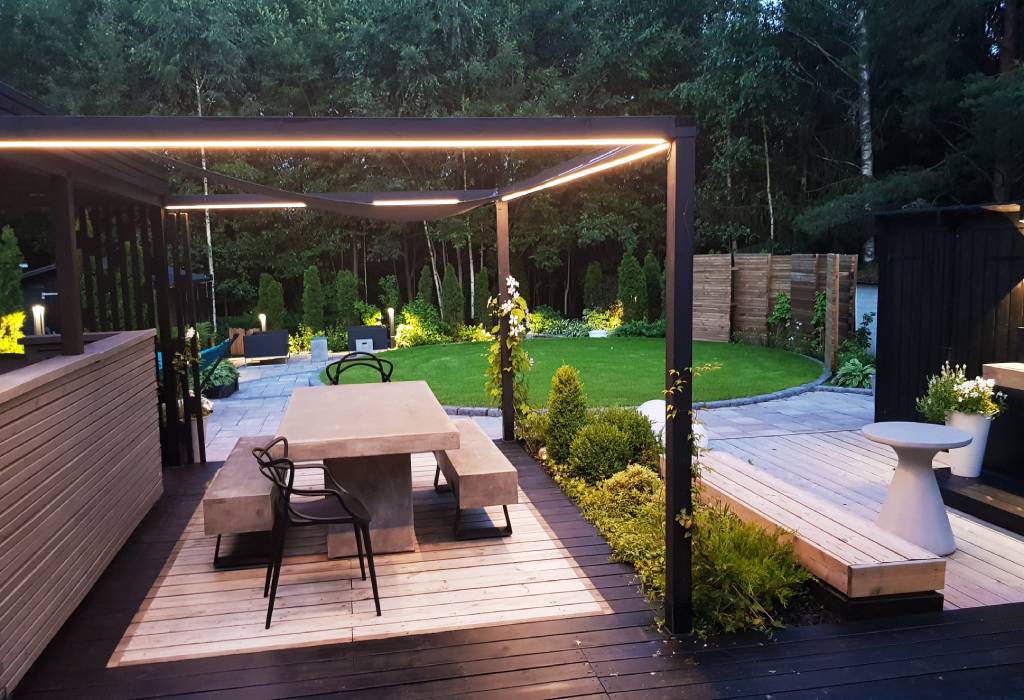Moderni takapihan patio, jossa on betoninen ruokapöytä ja mustat tuolit minimalistisen pergolan alla. Pergolaan on integroitu LED-valaistus luomaan tunnelmallista ilmettä. Aluetta ympäröivehreä kasvillisuus, yksi hoidettu nurmikko ja kivipolku, joka lisää luonnonläheistä viehätystä. Lisäksi pienenä yksityiskohtana löytyy valkoinen moderni istutuslaatikko elegantilla terassilla nauttimista varten. Tutustu LedStore.fi:n laajaan LED-valaistusvalikoimaan ja tuo samaa modernia eleganssia omaan pihaasi!