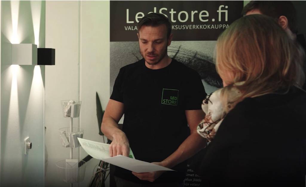 Mies mustassa LED Store -paidassa puhuu energisesti vaalean naisen kanssa, toinen henkilö kuuntelee lähellä; "LedStore.fi" kyltti ja kirkkaat LED-valot taustalla.