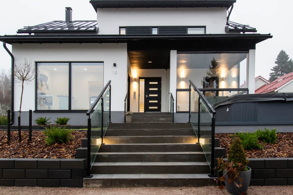 Moderni talo, jossa valkoinen julkisivu ja mustat aksentit; portaat lasikaiteilla LED-valoilla. Suuret ikkunat, minimaalinen maisemointi, pensaat ja naapuritalo.